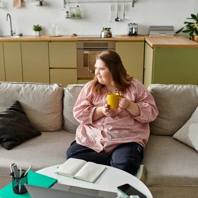 Young disabled women with down syndrome sipping coffee and sitting on sofa